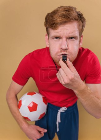 Foto de Joven hombre divertido entrenador de fútbol silbato mantener pelota de fútbol aislado en el estudio de fondo beige. Gente deporte humor concepto - Imagen libre de derechos