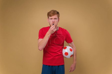Foto de Joven entrenador de fútbol silbato mantener pelota de fútbol aislado en estudio de fondo beige. Gente concepto deportivo - Imagen libre de derechos