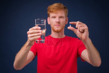Téléchargez les photos : Jeune homme sur fond bleu montre une pilule et un verre d'eau - en image libre de droit