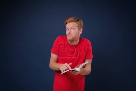 Photo for Young man study holding a book looking so confused and he does not understand anything, isolated on blue background - Royalty Free Image