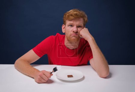 Téléchargez les photos : Drôle homme affamé mange un petit morceau de chocolat à partir d'une assiette à moitié vide. concept de faim et régime alimentaire - en image libre de droit