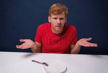 Téléchargez les photos : Jeune homme affamé à la table avec une assiette vide dans l'incrédulité. Le concept de jeûne - en image libre de droit