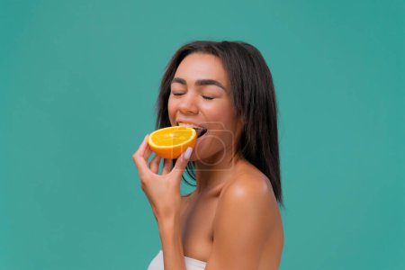 Foto de Mujer joven afroamericana mordiendo rebanada de naranja en fondo de estudio de color turquesa. El concepto de belleza y alimentación saludable. - Imagen libre de derechos