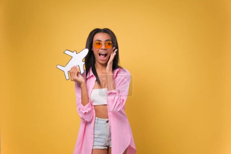 Photo for African American woman holds a paper plane in her hands. Airplane travel concept over isolated yellow background. - Royalty Free Image