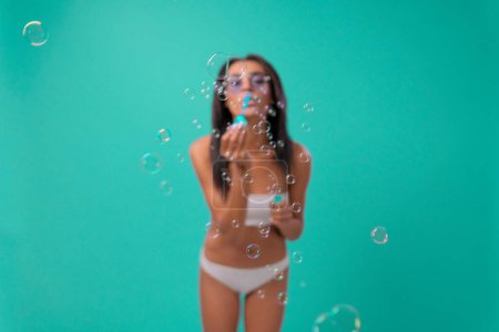 Téléchargez les photos : Sexy dame afro-américaine sur fond bleu en maillot de bain soufflant des bulles de savon. Hôtel d'été piscine mer repos soleil bronzage concept - en image libre de droit