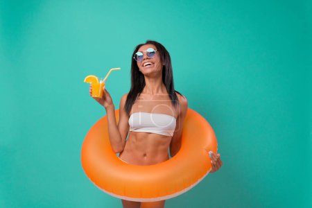 Foto de Mujer positiva con anillo inflable, celebración de cóctel tropical sobre fondo azul. Feliz dama afroamericana teniendo fiesta en la playa con flotador de la piscina y refrescante bebida de verano - Imagen libre de derechos