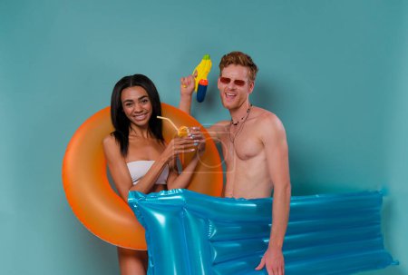 Téléchargez les photos : Fête de piscine amusant. Couple multiethnique avec anneaux gonflables s'amuser à tirer avec des pistolets à eau, fond studio bleu. Vacances d'été soleil de mer bronzage concept - en image libre de droit