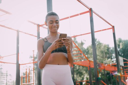 Téléchargez les photos : Souriant femme sportive afro-américaine utiliser et regarder sur smartphone sur le terrain de sport. Moderne femme saine et sportive mode de vie utilisation app. - en image libre de droit
