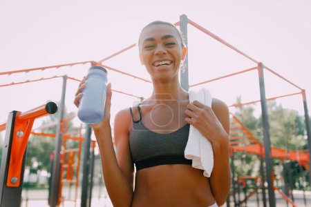 Foto de Atlética mujer africana en el patio de recreo con una botella de agua. Concepto de equilibrio hídrico. - Imagen libre de derechos