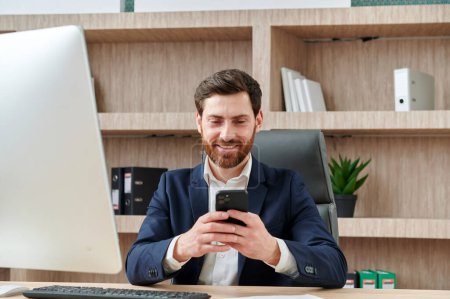 Foto de El hombre de negocios sonríe y escribe un mensaje en su teléfono inteligente en la oficina. Él está en un traje, lugar de trabajo en la oficina moderna y agradable. - Imagen libre de derechos
