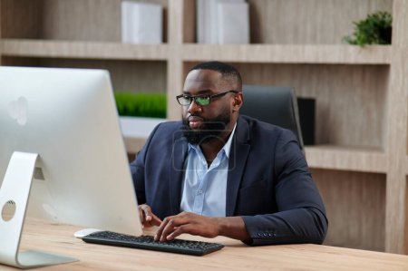 Téléchargez les photos : Homme d'affaires afro-américain concentré utilisant et tapant sur ordinateur pendant qu'il travaillait au bureau. Chef d'entreprise adulte assis au bureau. Concept d'homme moderne à succès. Idée de leadership et de gestion d'entreprise - en image libre de droit