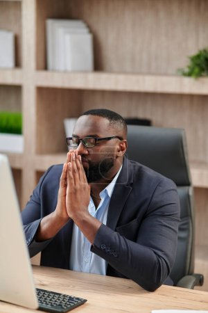 Foto de El tranquilo oficinista con barba medita con los ojos cerrados y las manos levantadas sobre un fondo azul. El hombre se calma. Afro-americano en traje de negocios - Imagen libre de derechos