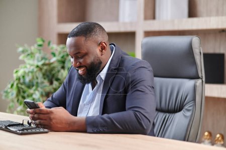 Téléchargez les photos : Heureux homme d'affaires afro-américain utilisant le téléphone mobile sur le lieu de travail SMS sms, homme souriant regardant smartphone navigation sur Internet, la technologie de bureau et la communication numérique - en image libre de droit