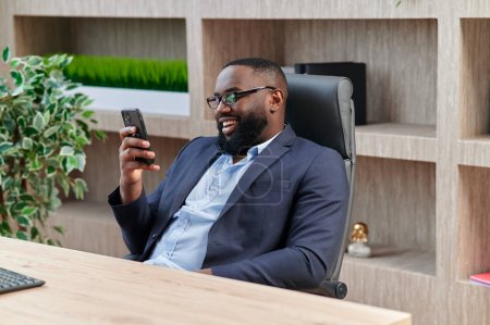 Foto de Feliz hombre de negocios afroamericano utilizando el teléfono móvil en el lugar de trabajo mensajes de texto SMS, hombre sonriente mirando el teléfono inteligente de navegación por Internet, la tecnología de oficina y la comunicación digital - Imagen libre de derechos