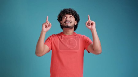 Photo for Young indian man wearing coral t-shirt isolated on a blue studio background. cheerful with a smile of face pointing with hand and finger up to the side with happy and natural expression on face - Royalty Free Image
