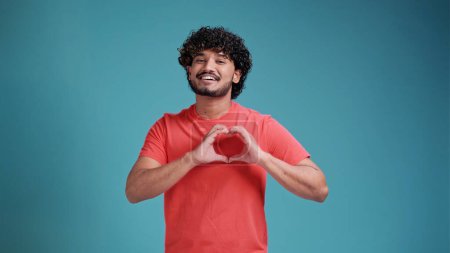 Photo for Handsome curly millennial hindu guy wearing coral t-shirt showing heart-shaped gesture over his chest and smiling at camera isolated on a blue studio background, copy space. Love, emotions concept. - Royalty Free Image