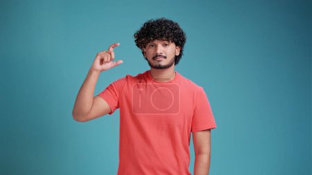 Téléchargez les photos : Homme indien millénaire déçu dans des lunettes montrant petite longueur de taille avec une pitié en corail t-shirt sur fond bleu studio, jeune homme regardant la caméra montrant la mesure avec les doigts - en image libre de droit