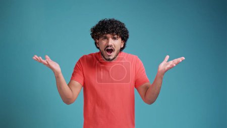 Photo for Shocked young bearded Indian man 20s years old says what. wears coral t-shirt on blue studio background. spreading hands shrugging shoulder standing questioned and unaware nothing to say. - Royalty Free Image