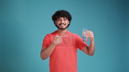 Foto de Sostenibilidad, consumismo y concepto ecológico - feliz hombre indio sonriente mostrando agua en vidrio reutilizable en camiseta de coral sobre fondo de estudio azul. - Imagen libre de derechos