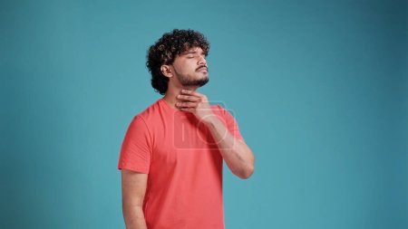 Téléchargez les photos : Homme indien barbu portant un t-shirt en corail touchant un cou douloureux, mal de gorge pour la grippe, la motte et l'infection sur fond bleu studio. - en image libre de droit