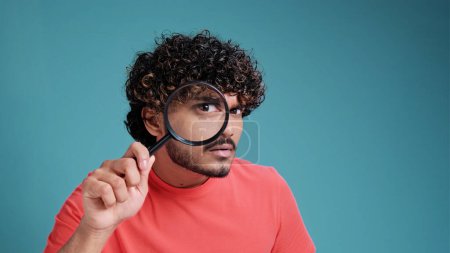 Téléchargez les photos : Homme drôle regardant à travers la loupe, cherchant ou enquêtant sur quelque chose, debout dans un pull orange sur fond turquoise - en image libre de droit