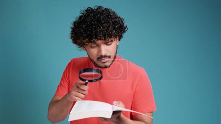 Téléchargez les photos : Homme indien drôle avec loupe lit livre en corail t-shirt sur fond bleu studio - en image libre de droit