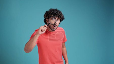 Téléchargez les photos : Homme drôle regardant à travers la loupe, cherchant ou enquêtant sur quelque chose, debout dans un pull orange sur fond turquoise - en image libre de droit