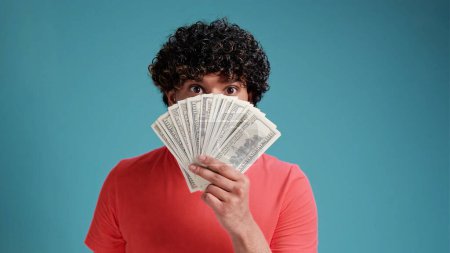 Photo for Smiling cheerful happy young bearded Indian man 20s years old wears coral shirt holding showing fan of cash money in dollar banknotes looking camera isolated on plain blue background studio portrait. - Royalty Free Image