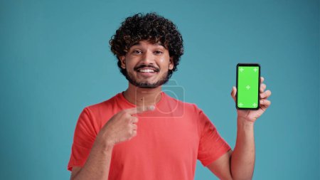Photo for Young indian man holding a phone with green screen, showing a blank chromakey screen in coral t-shirt on blue studio background. - Royalty Free Image