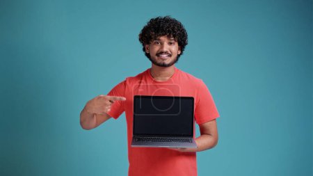 Photo for Portrait of positive joyful brunette Indian man with beard in white t-shirt standing holding laptop with blank screen and smiling at camera, internet advertising. studio shot isolated on blue studio background. - Royalty Free Image
