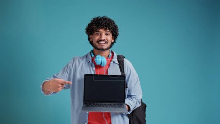 Photo for Handsome Indian guy on a blue background, studio photo, holding a laptop with a black screen in his hand and pointing at him with a hand gesture. - Royalty Free Image