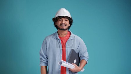 Foto de Concepto de personas y trabajo. El arquitecto positivo indio o árabe de la construcción, trabajador masculino, lleva un sombrero de trabajo que sostiene el plano sonríe alegremente a la cámara aislada sobre el fondo azul.. - Imagen libre de derechos