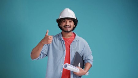 Photo for Young Indian or arabic architect man with helmet and holding blueprints and laptop over isolated on a blue background giving a thumbs up gesture. - Royalty Free Image