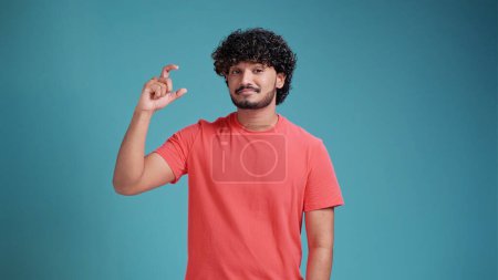 Photo for Disappointed millennial latin spanish man in glasses showing small size length with a pity in coral t-shirt on blue studio background, young guy looking at camera showing measurement with fingers - Royalty Free Image