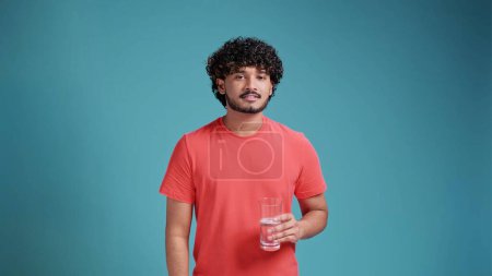 Foto de Sostenibilidad, consumismo y concepto ecológico - feliz hombre español latino sonriente mostrando agua en vidrio reutilizable en camiseta de coral sobre fondo azul estudio. - Imagen libre de derechos