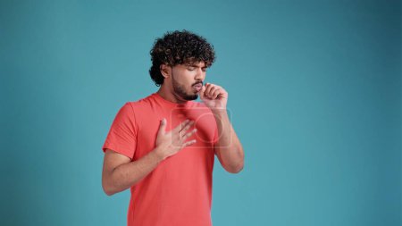 Photo for Young latin spanish man in coral t-shirt on blue studio background, scarf sneezing or coughing covering mouth with hands. - Royalty Free Image