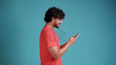 Photo for Dreamful marvelous fun young bearded Indian man 20s years old hold in hand use mobile cell phone typing browsing chatting send sms in coral t-shirt on blue studio background studio portrait - Royalty Free Image