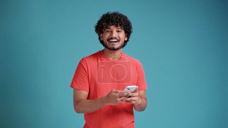 Téléchargez les photos : Dreamful merveilleux amusant jeune homme indien barbu 20 ans tenir à la main utiliser téléphone portable dactylographier navigation chat envoyer sms en corail t-shirt sur fond bleu studio portrait studio - en image libre de droit