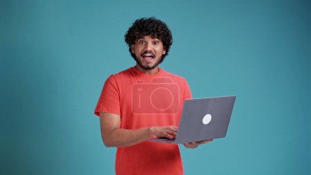 Happy indian man winner wearing earbud winning online raising fist in yes gesture watching game on laptop screaming about goal win celebrating great prize isolated on yellow background.