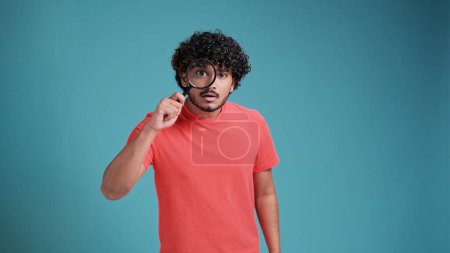 Foto de Divertido joven latino español chico con una lupa en las manos mira hacia fuera o busca algo, vestido de casual, sobre un fondo de estudio azul - Imagen libre de derechos