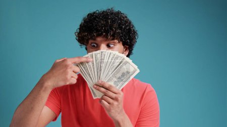 Photo for Smiling cheerful happy young bearded latin spanish man 20s years old showing fan of cash money in dollar banknotes looking camera isolated on plain blue background studio portrait. - Royalty Free Image