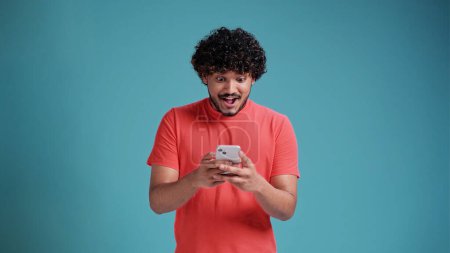 Foto de Joven latino mira encantado en el teléfono inteligente y dice wow. Vestido de casual en un fondo de estudio azul - Imagen libre de derechos