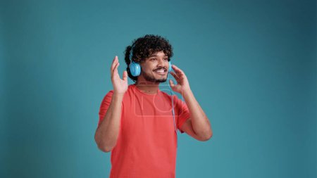 Téléchargez les photos : Jeune souriant heureux heureux homme latin ou arabe 20s avec écouteurs écouter de la musique s'amuser sur fond bleu studio. Concept de style de vie - en image libre de droit