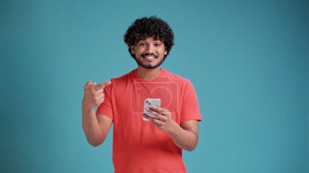 Foto de Conduce a un hombre en una camiseta de coral señala un dedo a un teléfono inteligente y sonríe mientras está de pie sobre un fondo de estudio azul - Imagen libre de derechos
