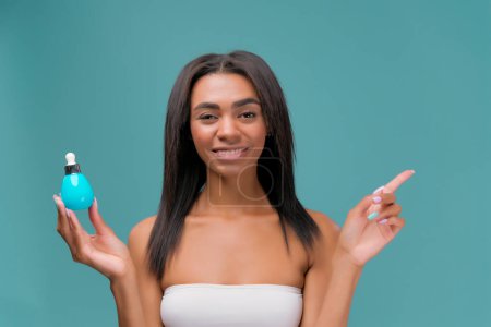 Téléchargez les photos : Souriant heureux belle jeune femme afro-américaine avec sérum hyaluronique avec pipette pointant le doigt sur le côté dans un espace vide, isolé sur fond bleu - en image libre de droit