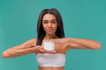 Foto de Mujer joven afroamericana medio desnuda mostrando un frasco blanco de crema. Concepto de belleza sobre fondo de estudio azul - Imagen libre de derechos