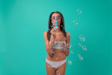 Photo for Multiethnic sexy woman on blue background in swimsuit blowing soap bubbles. Summer hotel pool sea rest sun tan concept - Royalty Free Image