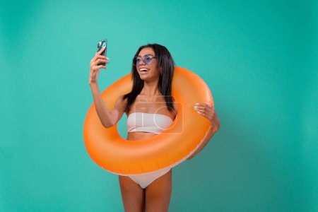 Foto de Mujer joven afroamericana en traje de baño y con un anillo de piscina mira en un teléfono inteligente y sonríe o se toma una selfie en un fondo de estudio azul - Imagen libre de derechos