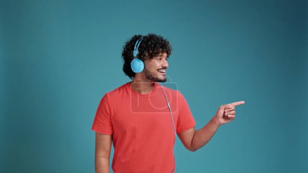 Téléchargez les photos : Swarthy man écoute de la musique à l'aide d'écouteurs en t-shirt corail sur fond bleu studio. Très heureux pointant avec la main sur le côté - en image libre de droit