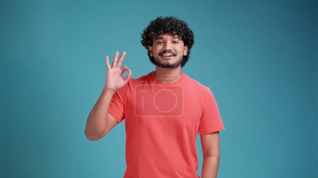 Photo for Everything is OK positive latin spanish man showing OK sign . close up photo. gesture concept, in basic coral t-shirt isolated on a blue studio background. - Royalty Free Image